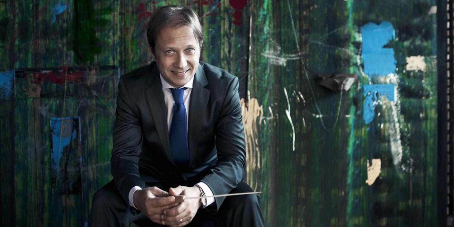 A conductor in a dark suit and blue tie sits smiling with his baton in hand, in front of a large green abstract painting