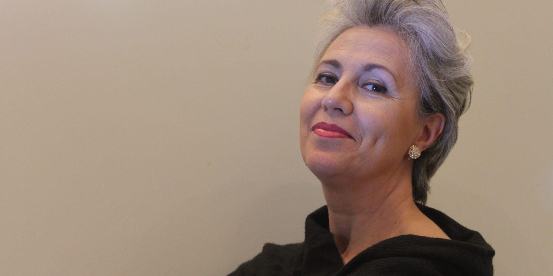 Contralto con capelli corti grigio-argento, labbra rosse e abito nero, ritratta di tre quarti con lo sguardo sereno rivolto verso l'alto su sfondo beige neutro
