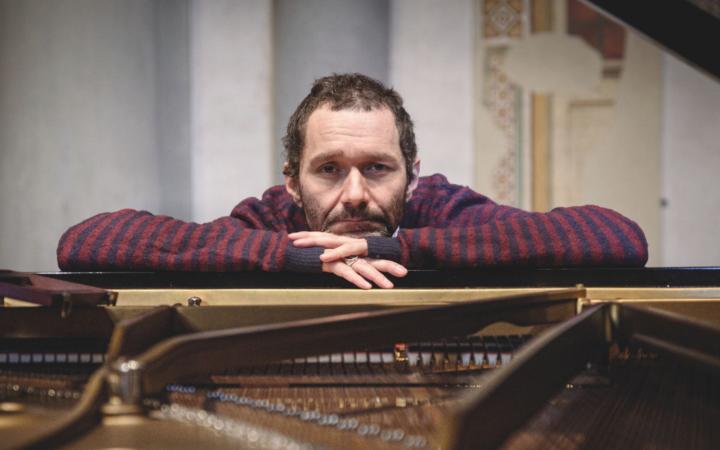Ritratto frontale del musicista Davide “Boosta” Dileo, appoggiato al bordo di un pianoforte a coda aperto.