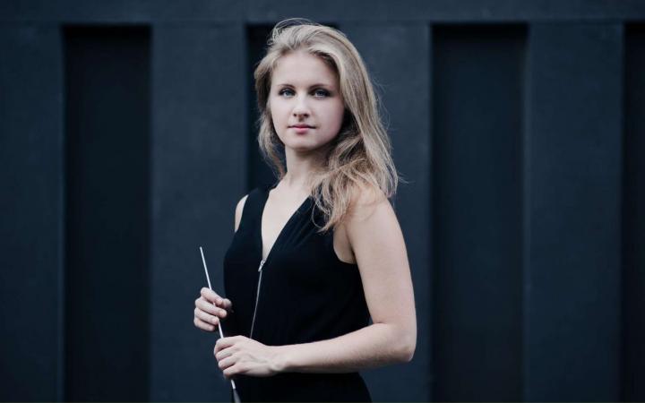 Una giovane direttrice d'orchestra bionda in abito nero tiene la bacchetta davanti a sé, su sfondo di pannelli grigio scuro