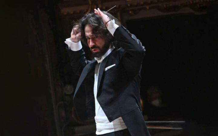 Un direttore d'orchestra con capelli lunghi scuri e barba, in frac, dirige con intensa energia tenendo la bacchetta alzata sopra la testa