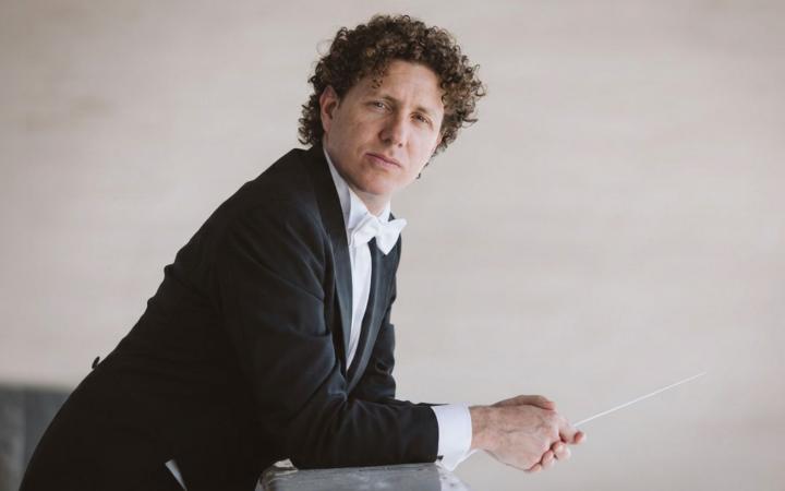 Un direttore d'orchestra con capelli ricci castani in frac con cravatta bianca, appoggiato a una superficie, tiene la bacchetta con sguardo pensieroso verso l'obiettivo