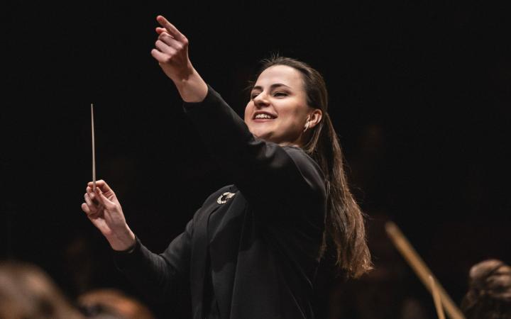 Una giovane direttrice d'orchestra bruna in abito nero dirige con sorriso radioso, bacchetta in mano e braccio sinistro alzato, davanti all'orchestra in penombra