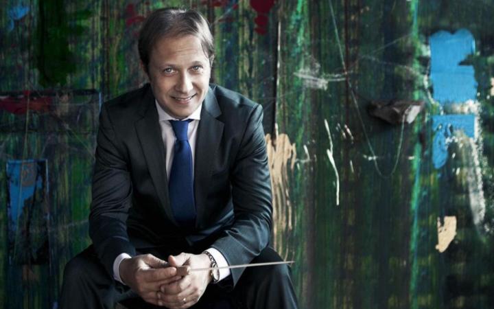 A conductor in a dark suit and blue tie sits smiling with his baton in hand, in front of a large green abstract painting