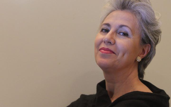 Contralto con capelli corti grigio-argento, labbra rosse e abito nero, ritratta di tre quarti con lo sguardo sereno rivolto verso l'alto su sfondo beige neutro