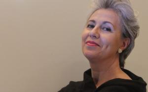 Contralto con capelli corti grigio-argento, labbra rosse e abito nero, ritratta di tre quarti con lo sguardo sereno rivolto verso l'alto su sfondo beige neutro